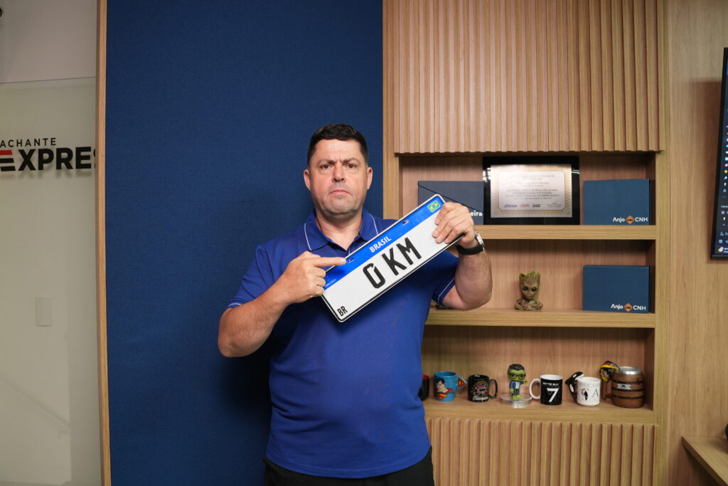 Homem segurando uma placa Mercosul com a inscrição "0KM" e expressão séria, destacando o direito de fazer a documentação do veículo fora da concessionária.