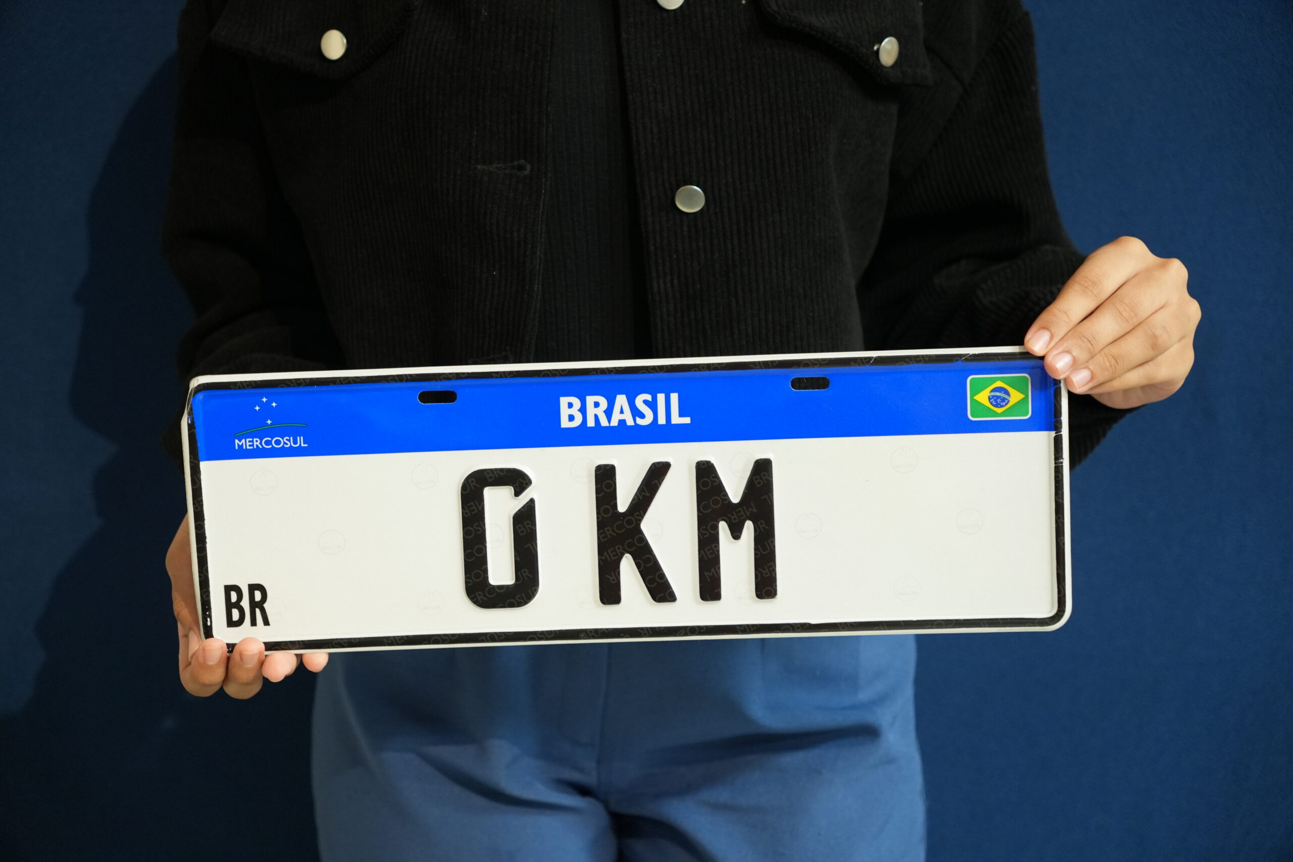 Pessoa segurando uma placa Mercosul com a inscrição "0KM", simbolizando o emplacamento de veículo novo fora da concessionária.